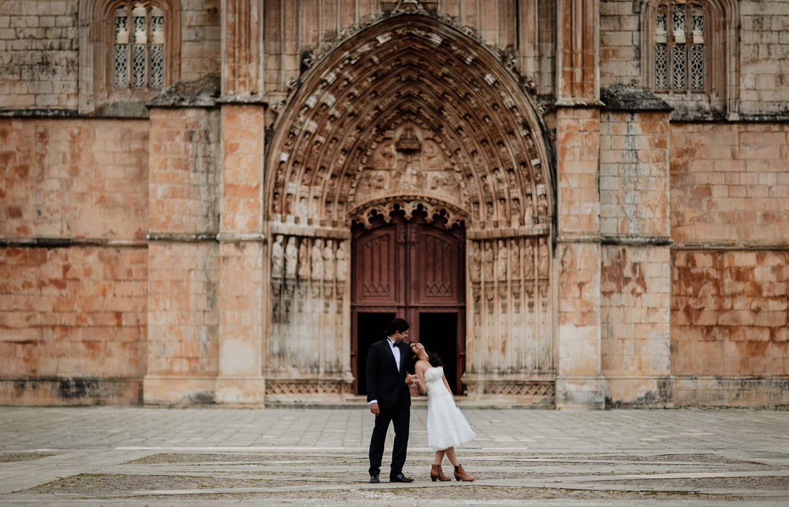 A photograph of a bride and groom playfully posing in-front of a grand cathedral door