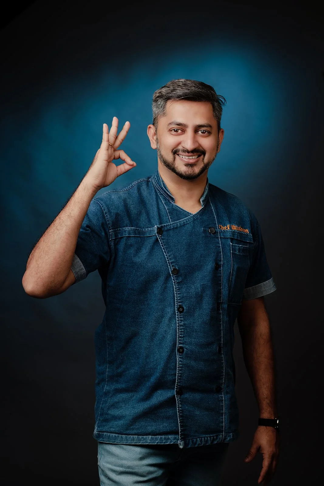 A corporate headshot photograph of a chef with a blue shirt on a blue backdrop
