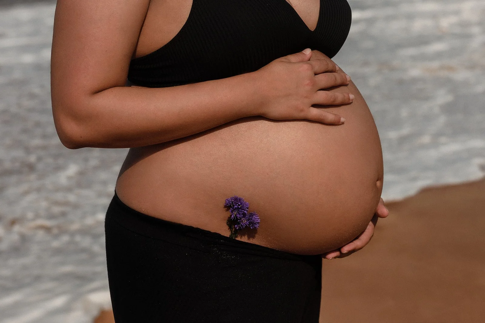 A maternity photograph of a lady's belly with a beautiful purple flower tucked under and the waves crashing behind her