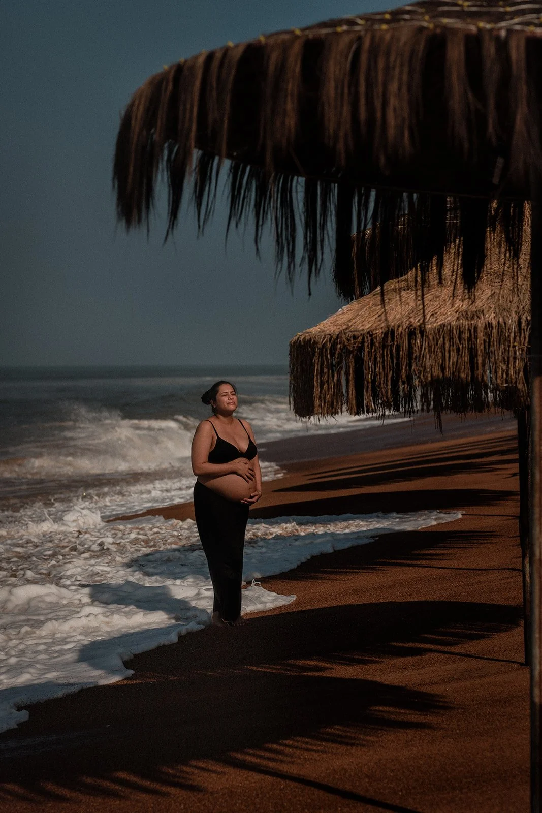 A maternity photograph of a lady basking in the light on the beach with a few shacks around her and her holding her belly 