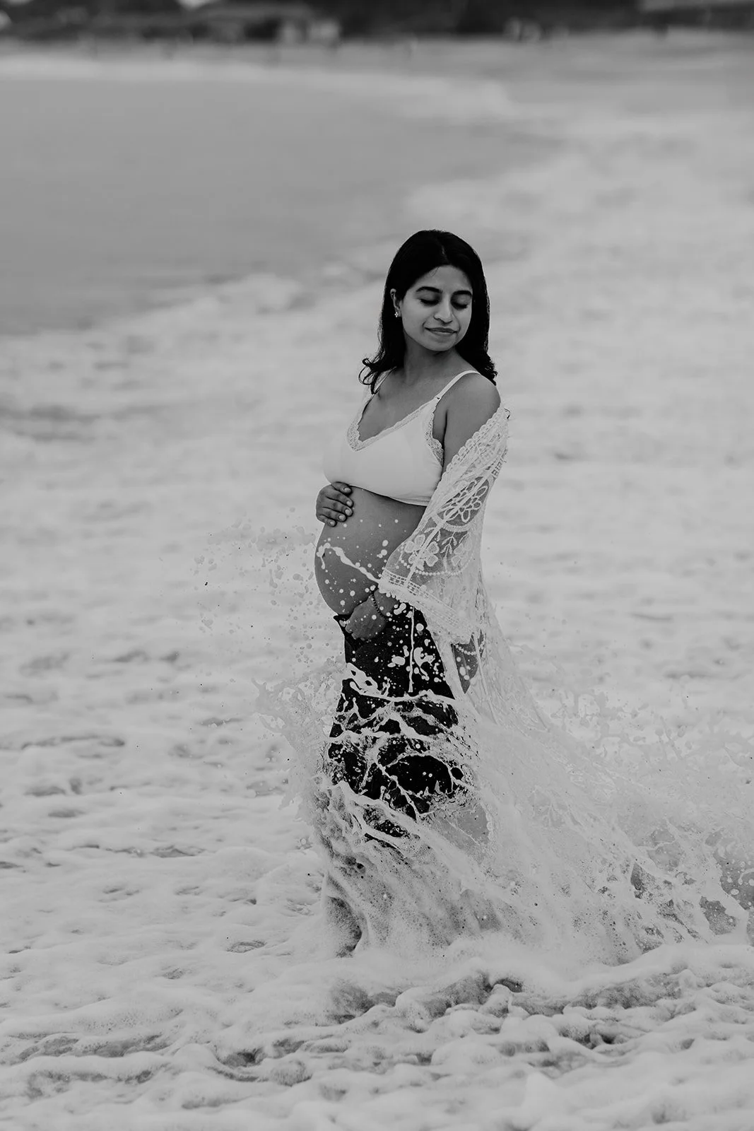 A Black and white maternity photograph of a lady in the waves holding her pregnant belly 