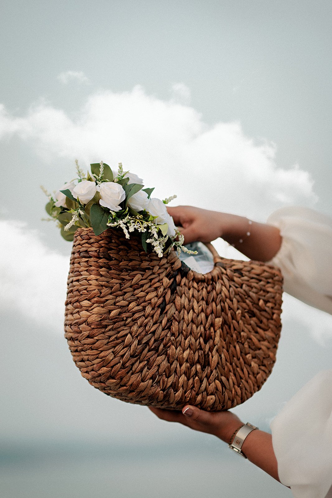 the bride to be holding a woven handbag with white flowers and green leaves against a cloudy sky at a bachelorette party