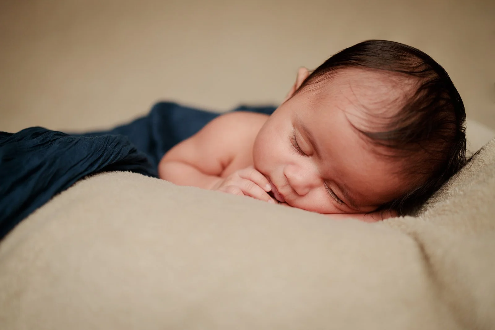 A new born photograph of a child sleeping on a cream colored backdrop with a blue cloth around him 