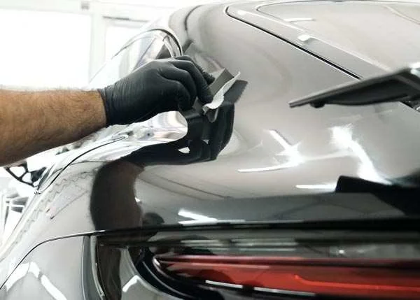 Person polishing or repairing the rear of a silver or dark-colored car, using a cloth or sponge, and wearing a black glove.