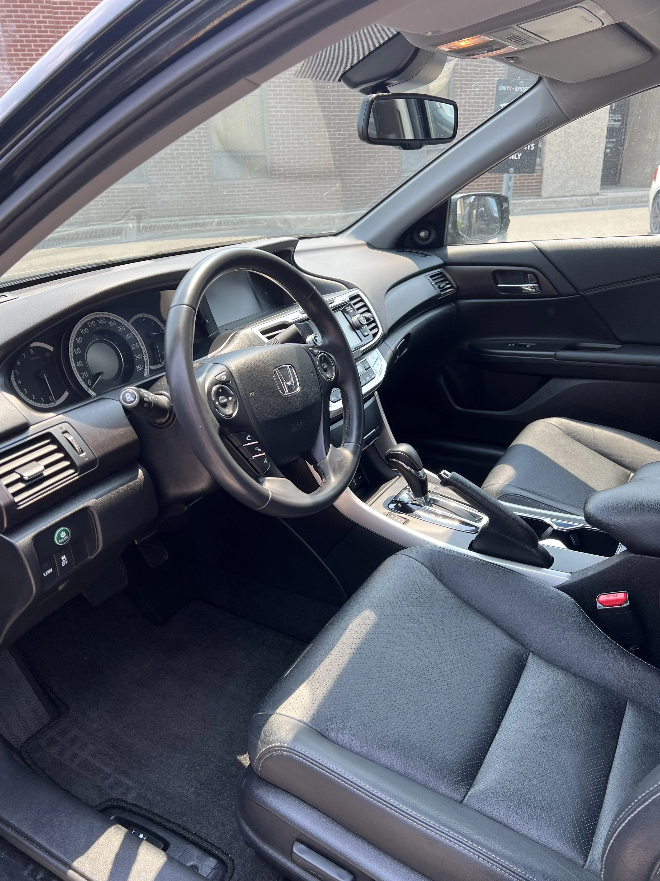 Interior of a Honda car showing the dashboard, steering wheel, front seats, and center console, with a view through the front windshield and side mirror of a brick building outside.
