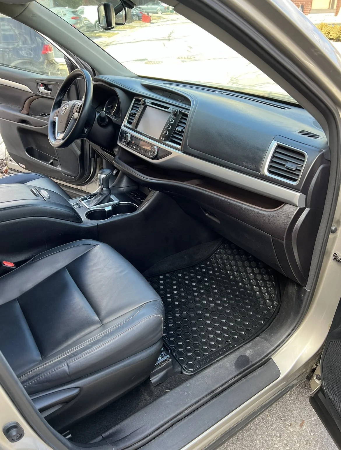 Interior of a vehicle showing the dashboard, steering wheel, gear shift, and front seats with black leather upholstery. The vehicle is parked outdoors, with other cars visible through the windows.