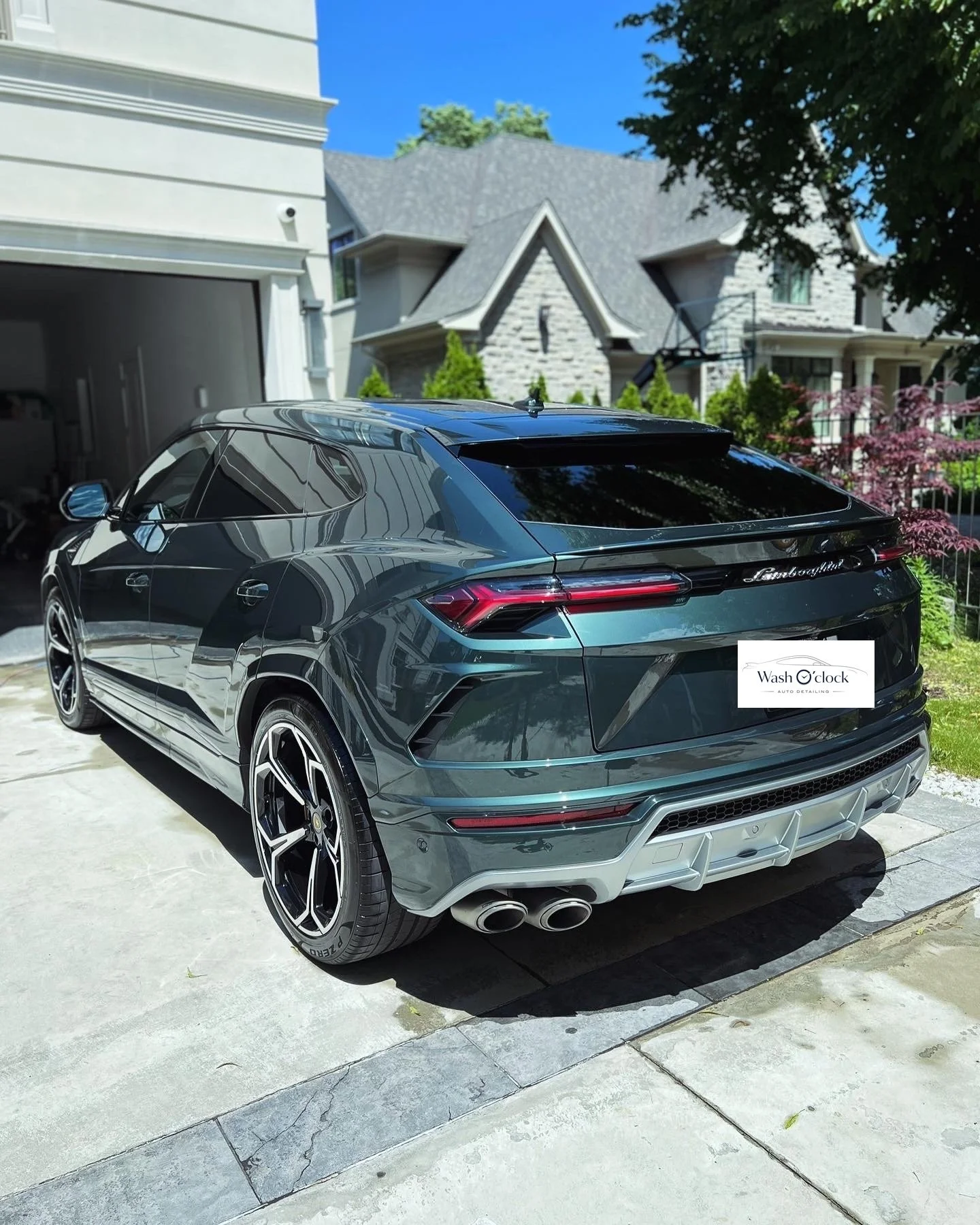 A sleek, black Lamborghini SUV parked in a driveway in front of a modern house with a gray roof and stone exterior. The driveway is concrete, and there are shrubs and trees around. The sky is clear and blue.