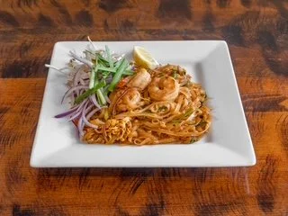 Plate of fried noodles topped with shrimp, garnished with lime, green onions, and sliced red onions on a wooden table.