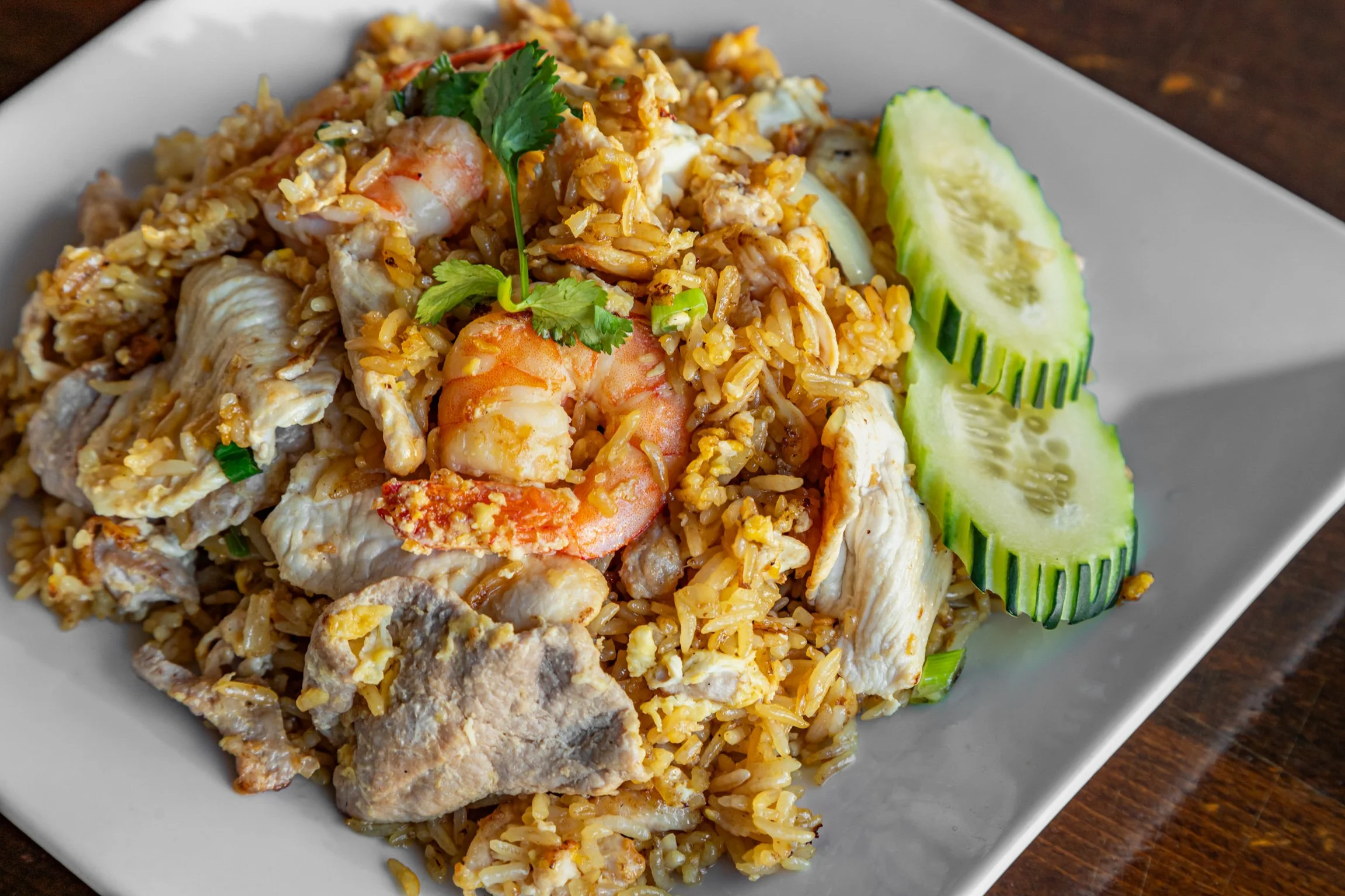 Plate of fried rice with shrimp, chicken, and vegetables garnished with cilantro, served with sliced cucumber on a white dish.