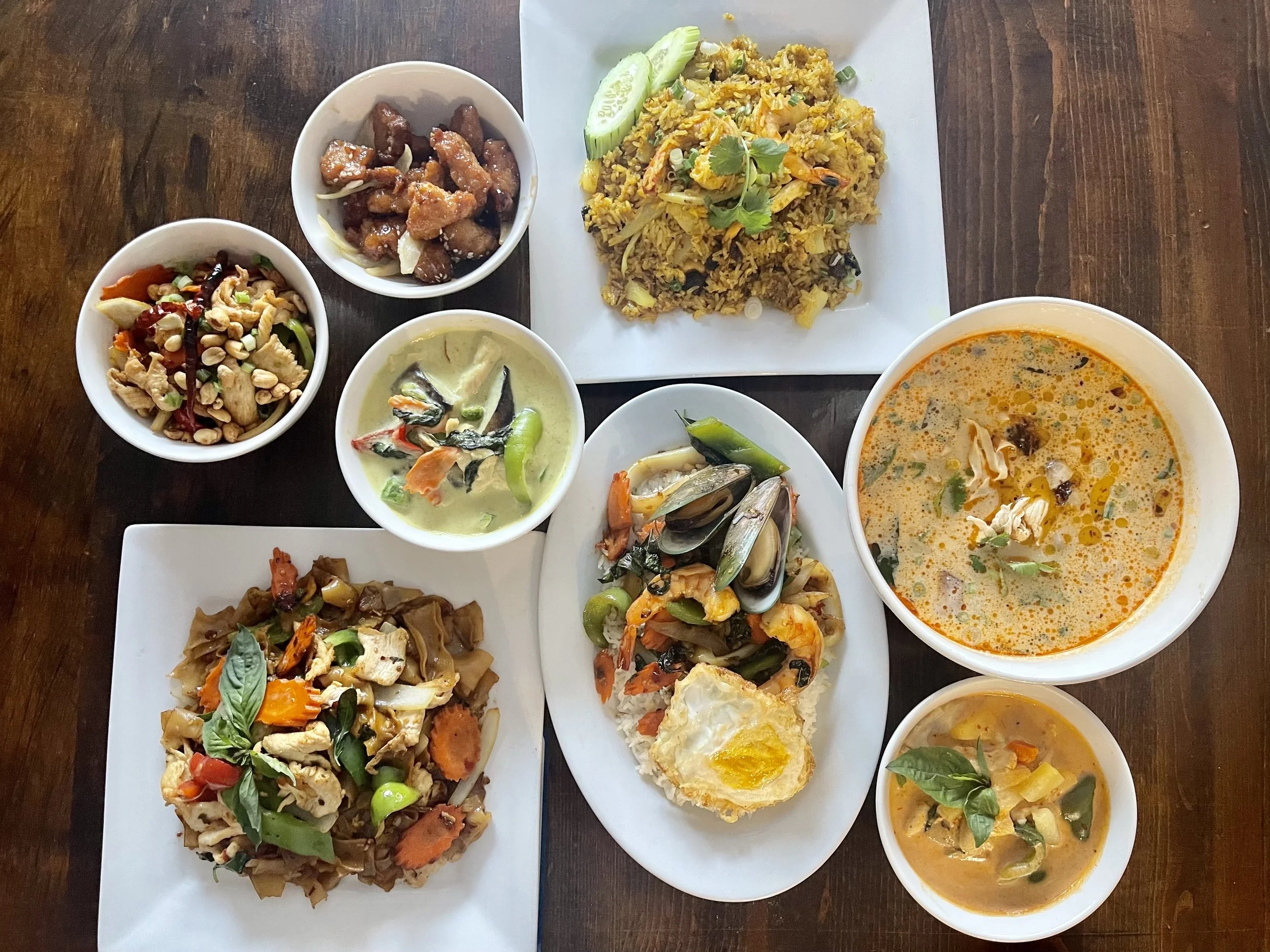 Assorted Thai dishes including fried rice, seafood, vegetable stir-fry, curry, and small side dishes, served on white and square plates, with cucumbers on the side, all placed on a dark wooden table.