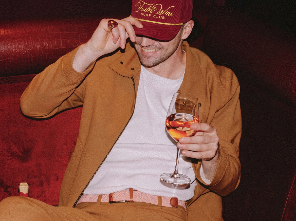 Man wearing a red Toilet Wine Surf Club cap with gold embroidery and a tan jacket, holding a glass of rosé wine.