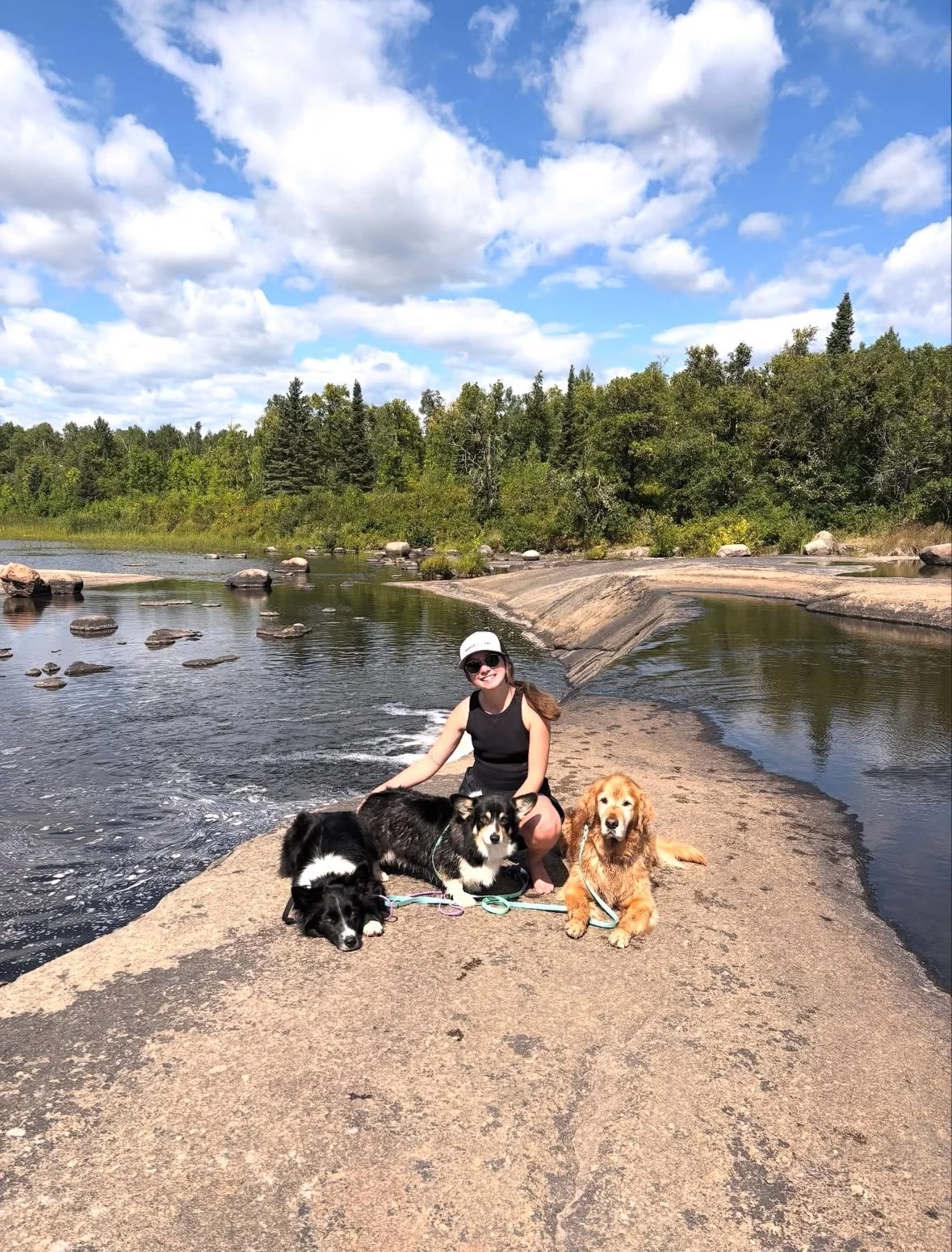 A woman sitting with four dogs on a large flat rock beside a river, surrounded by lush greenery and trees, under a partly cloudy sky.
