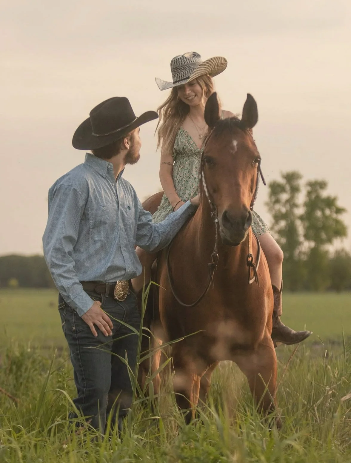 A man and woman with cowboy hats and Western-style clothing with a sheriff's badge on his belt, standing in a grassy field, with the woman sitting on a brown horse wearing black ears, during sunset.