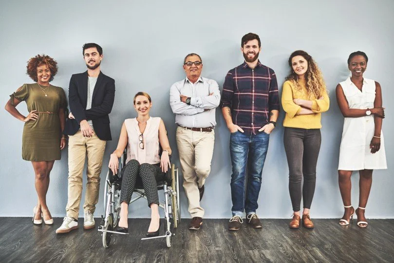 Group of seven diverse adults standing against a plain wall, one woman in a wheelchair, smiling.