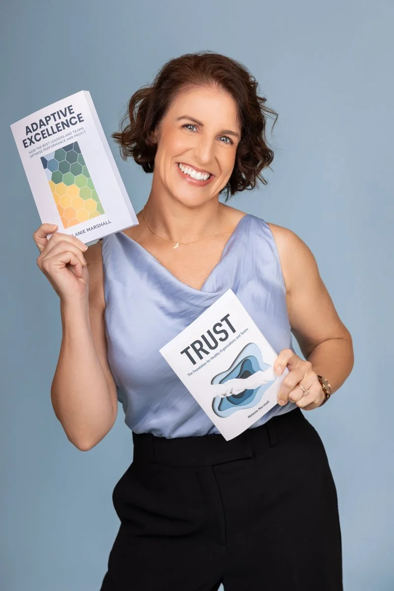 Melanie Marshall, wearing a light blue sleeveless blouse and black pants, smiling and holding two books she's authored titled 'Adaptive Excellence' and 'Trust'.