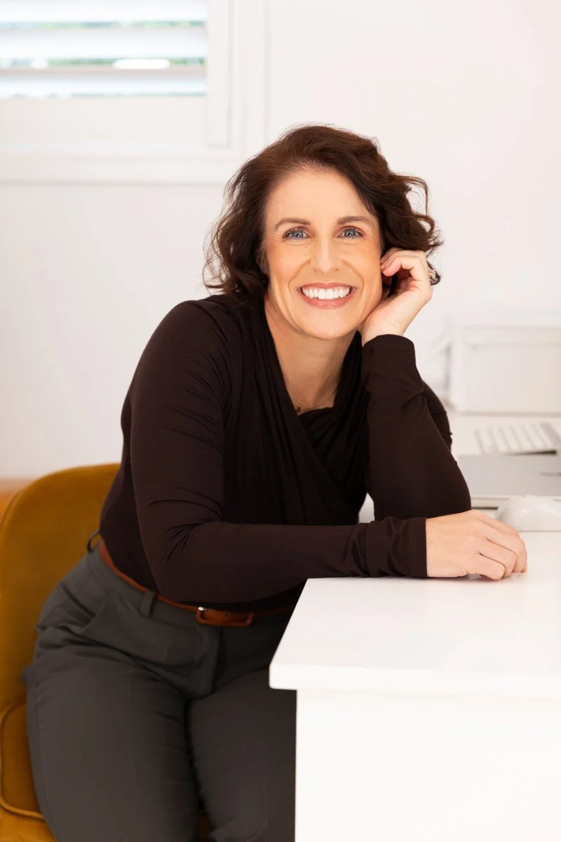 Melanie Marshall with brown hair and blue eyes sitting at a white desk, smiling, indoors with a white wall and window in the background.