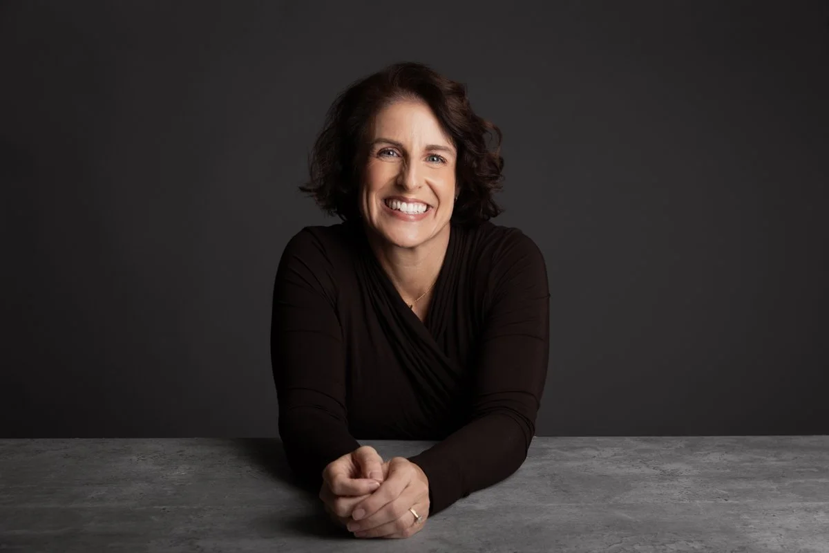 Melanie Marshall smiling and leaning on a table against a dark gray background.