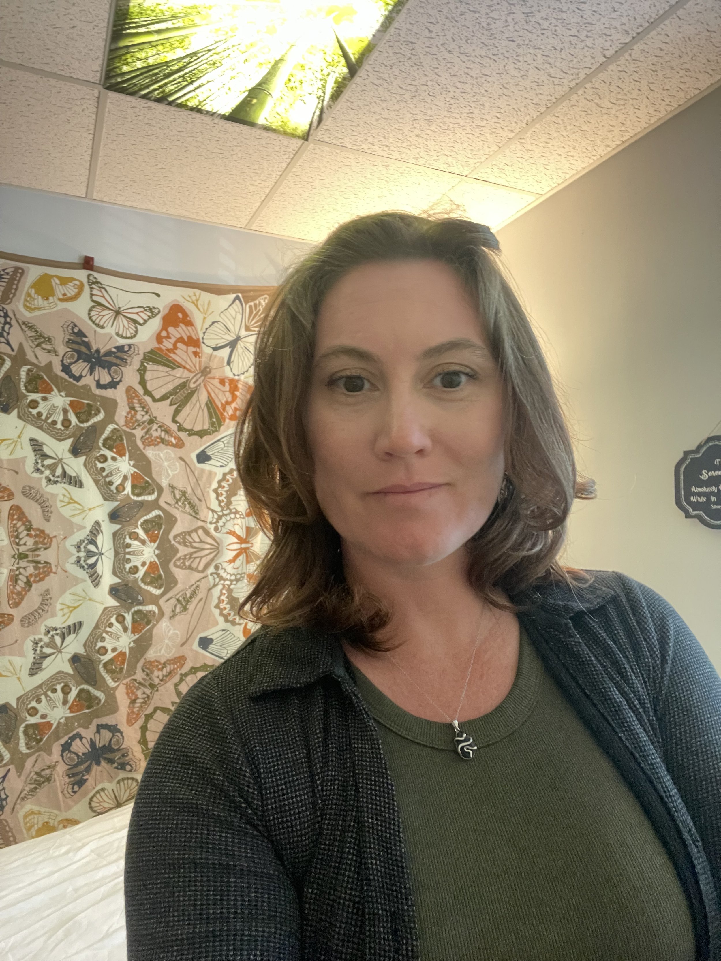 A woman with shoulder-length brown hair taking a selfie in a room with a butterfly-themed tapestry, a ceiling with an image of bamboo, and soft lighting.