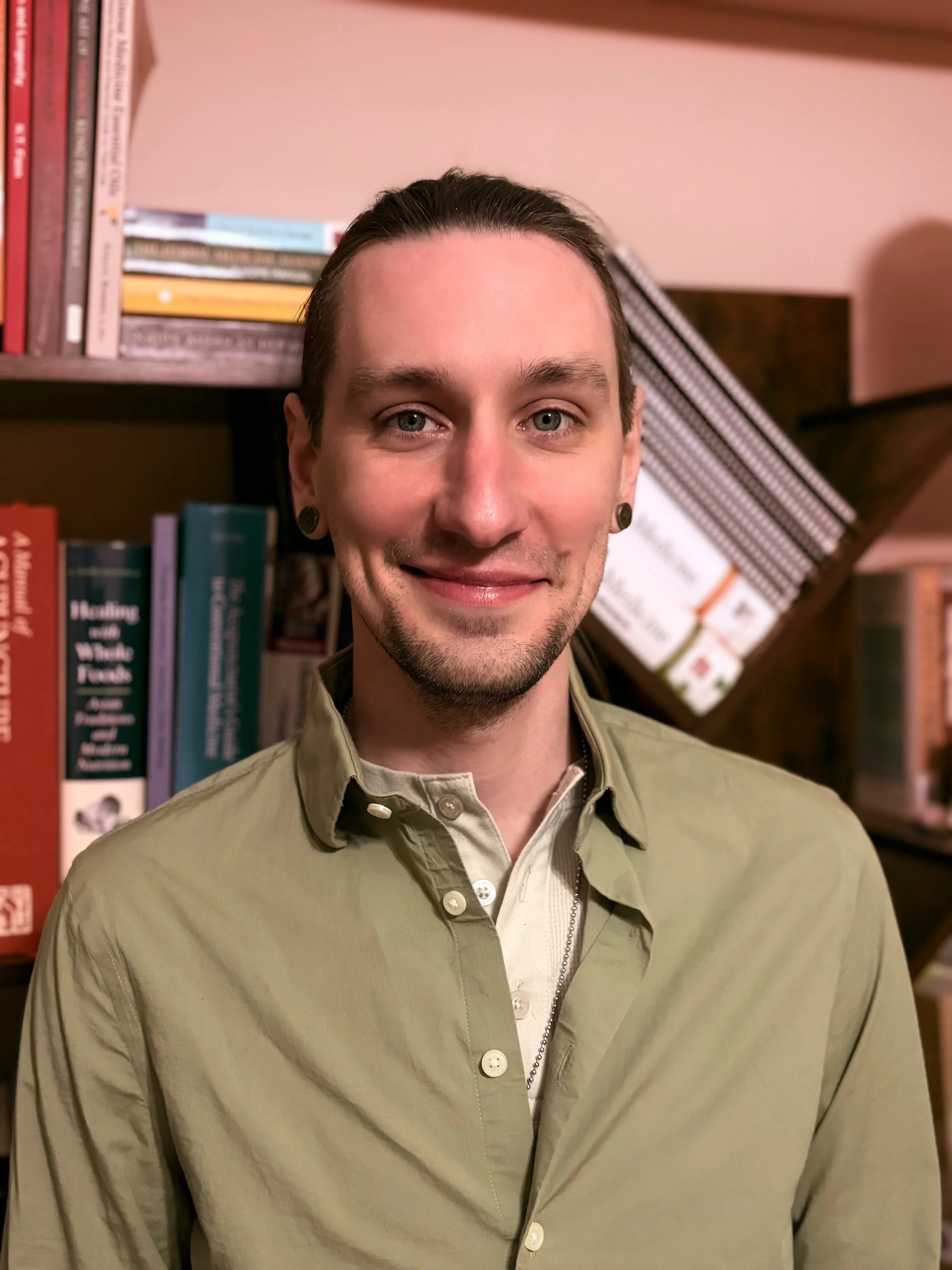 A young man with dark hair, earrings, and a beard wearing a beige button-up shirt, standing in front of a bookshelf filled with books.