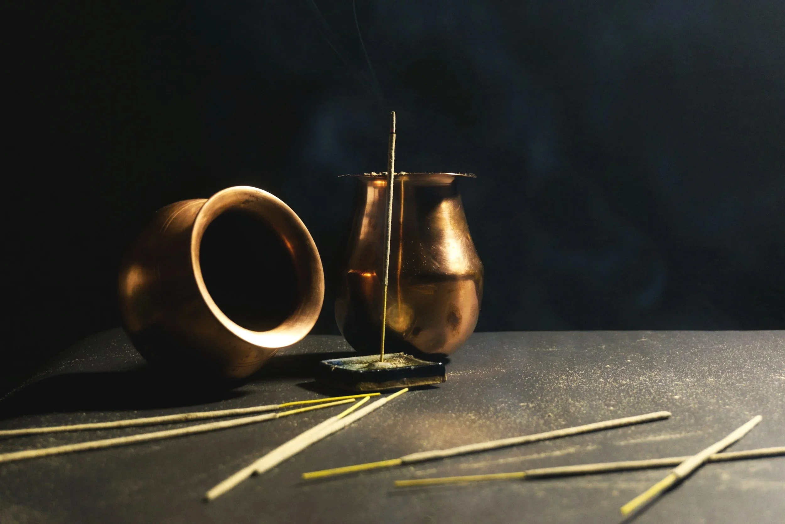 Two metallic vases with a dark background; one vase is lying on its side, and the other is upright. Several incense sticks are on the surface, with two burning sticks emitting smoke.
