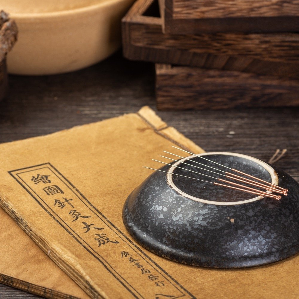 Traditional acupuncture needles resting on a black ceramic dish, placed on an aged newspaper with Asian characters, on a wooden surface.