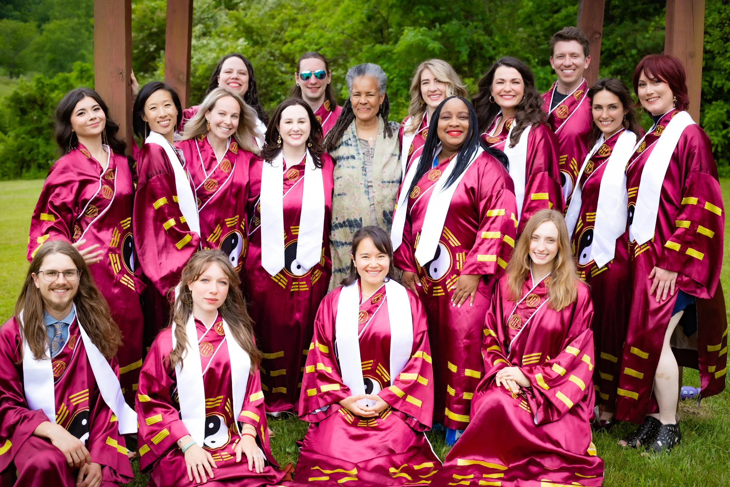 Group of graduates wearing maroon robes with yin-yang symbols, some with white stoles, posing outdoors on grass near a wooden arch with greenery in the background.