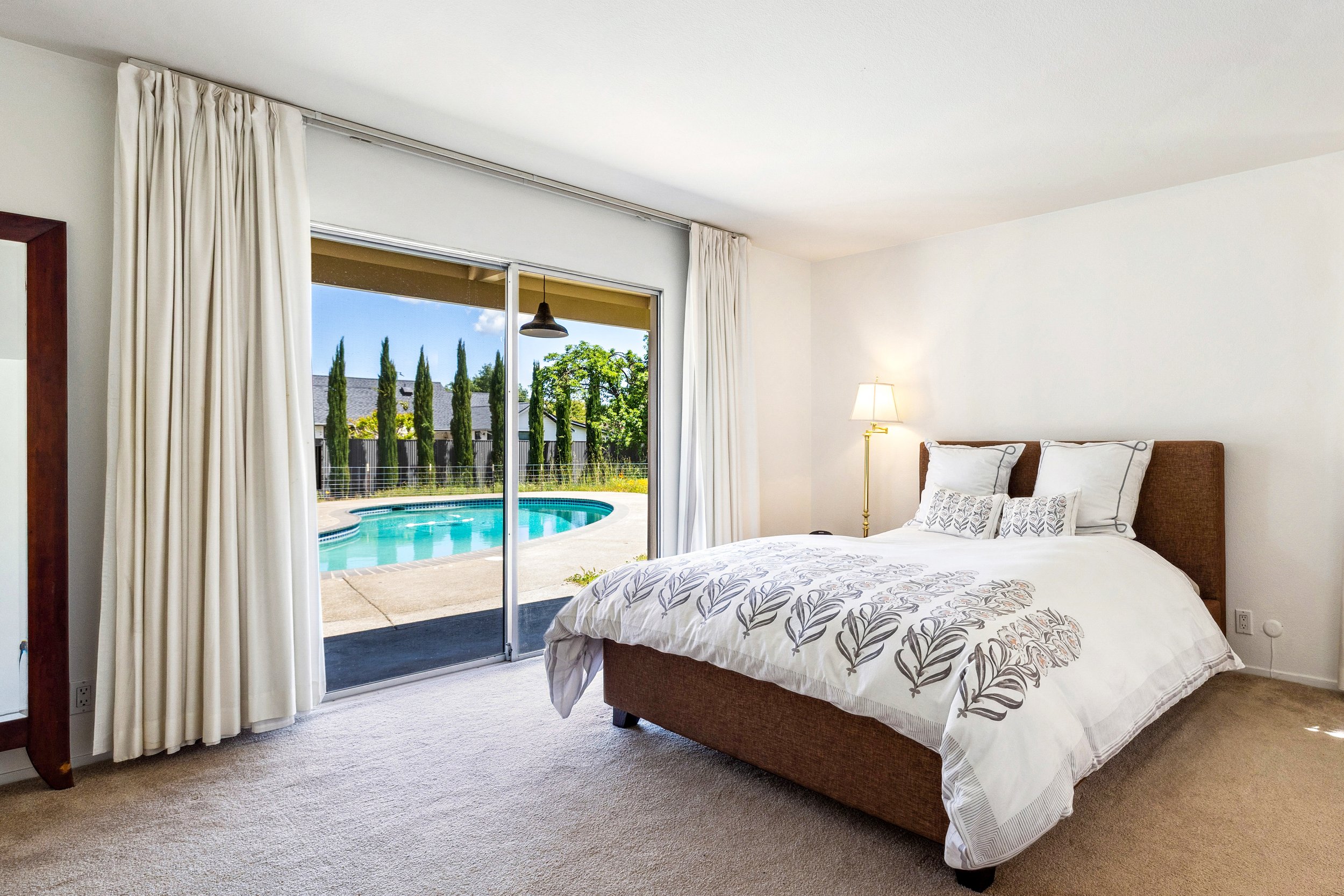 Bedroom with a large bed, white bedding with leaf pattern, pillows, a floor lamp, and sliding glass doors leading to a backyard with a swimming pool and tall trees.