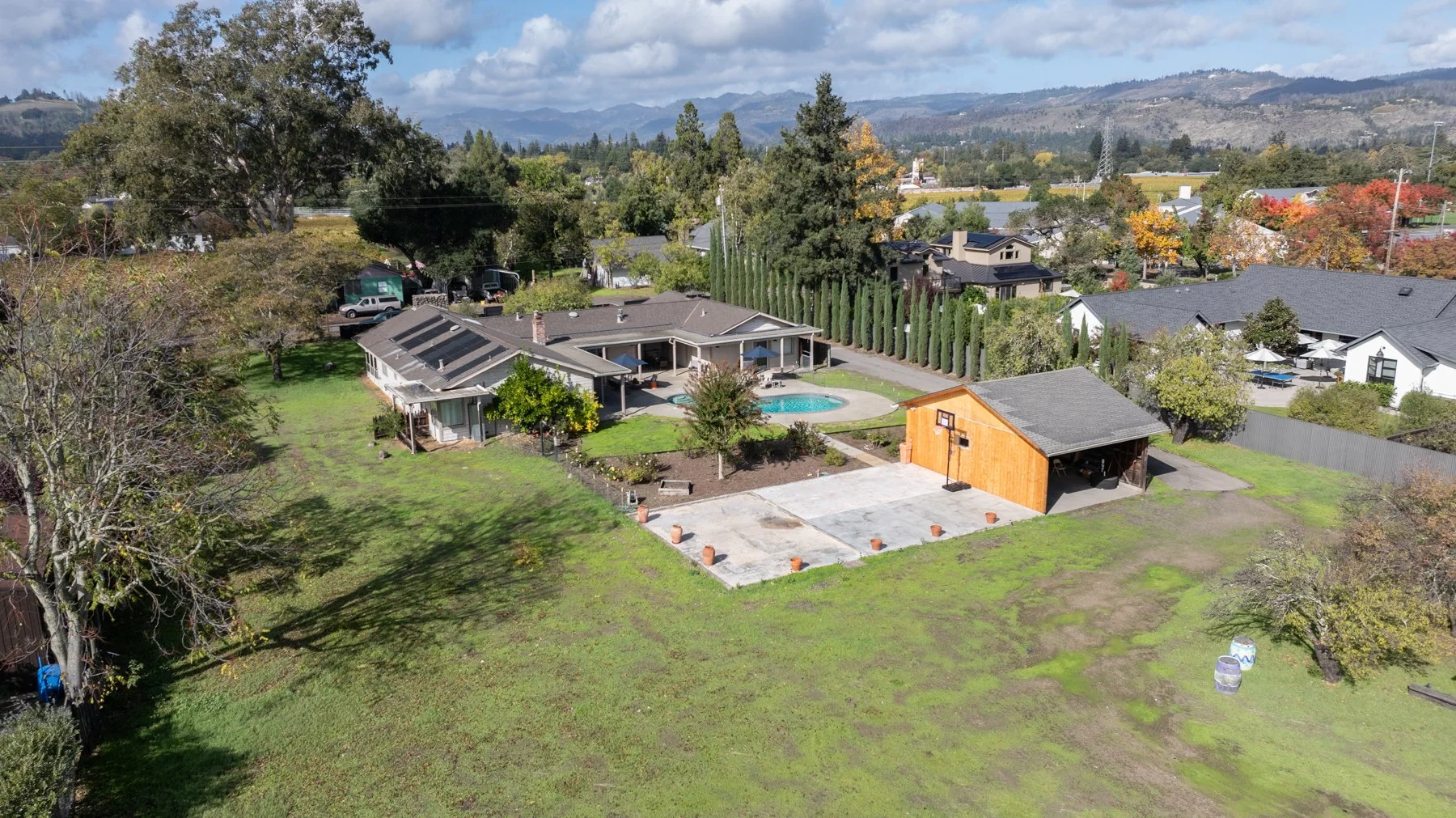 Aerial view of a residential backyard with a swimming pool, a covered patio, a detached wooden structure, a concrete driveway with potted plants, and surrounding houses with trees and hills in the background.