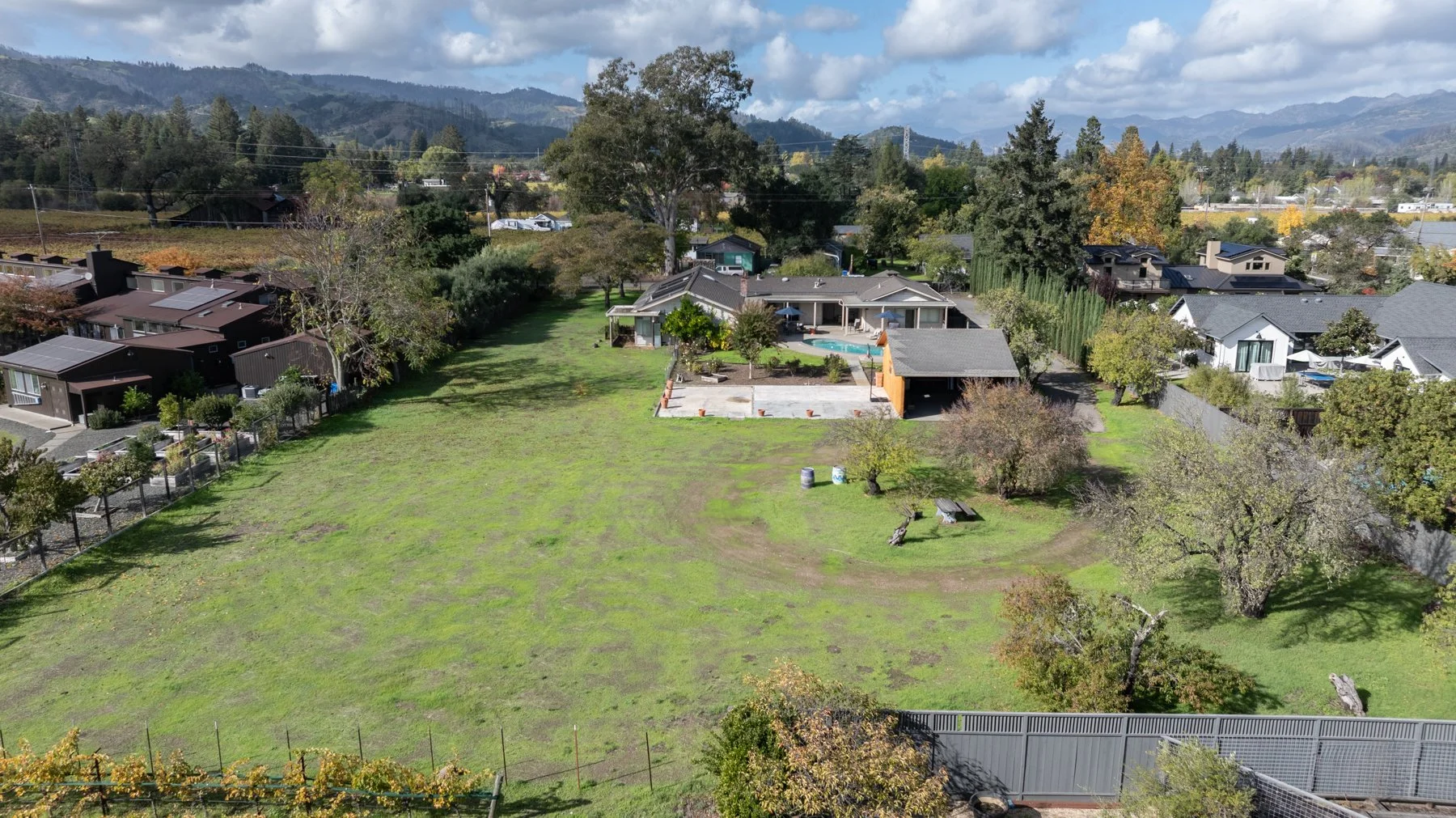 A large backyard with a house, a pool, trees, and a field, surrounded by neighboring houses and mountains in the background.