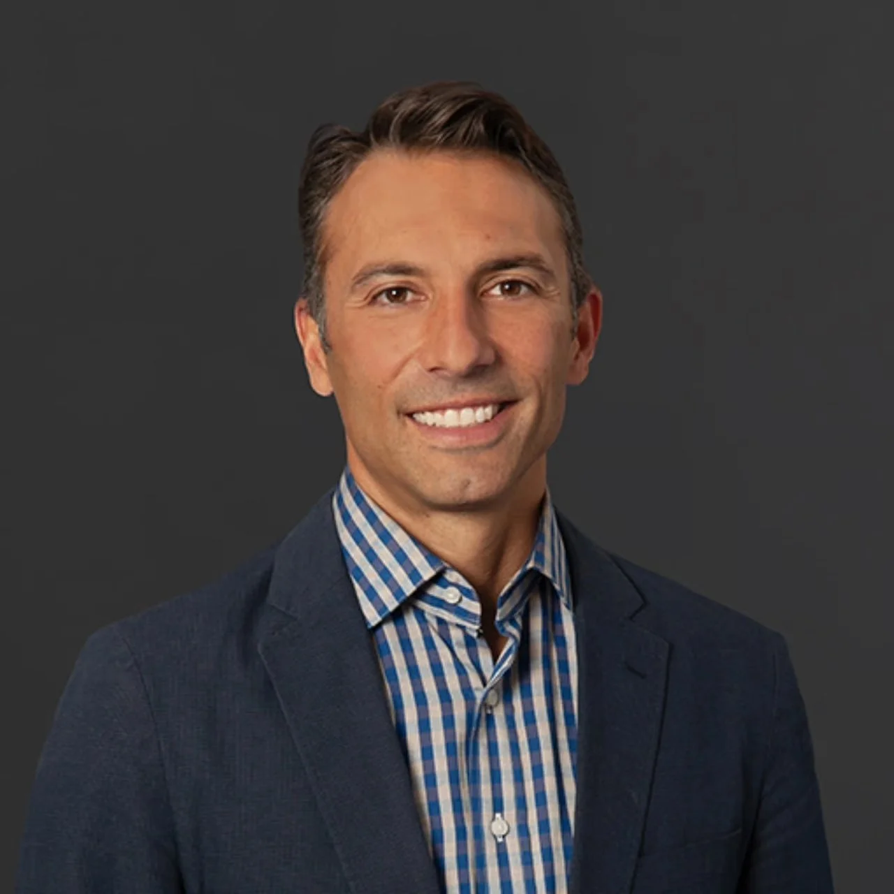 Professional man smiling, wearing a bright blue suit and a blue and white checkered shirt, with a dark gray background.