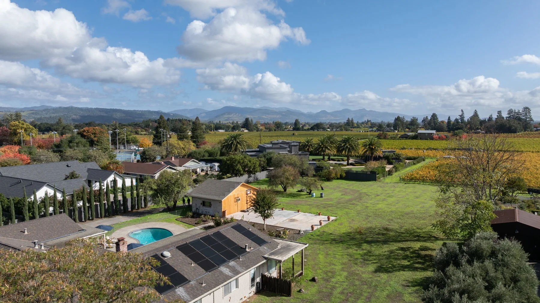 A scenic aerial view of a suburban neighborhood with houses, trees, a swimming pool, and a large grassy yard, with mountains in the background under a partly cloudy sky.