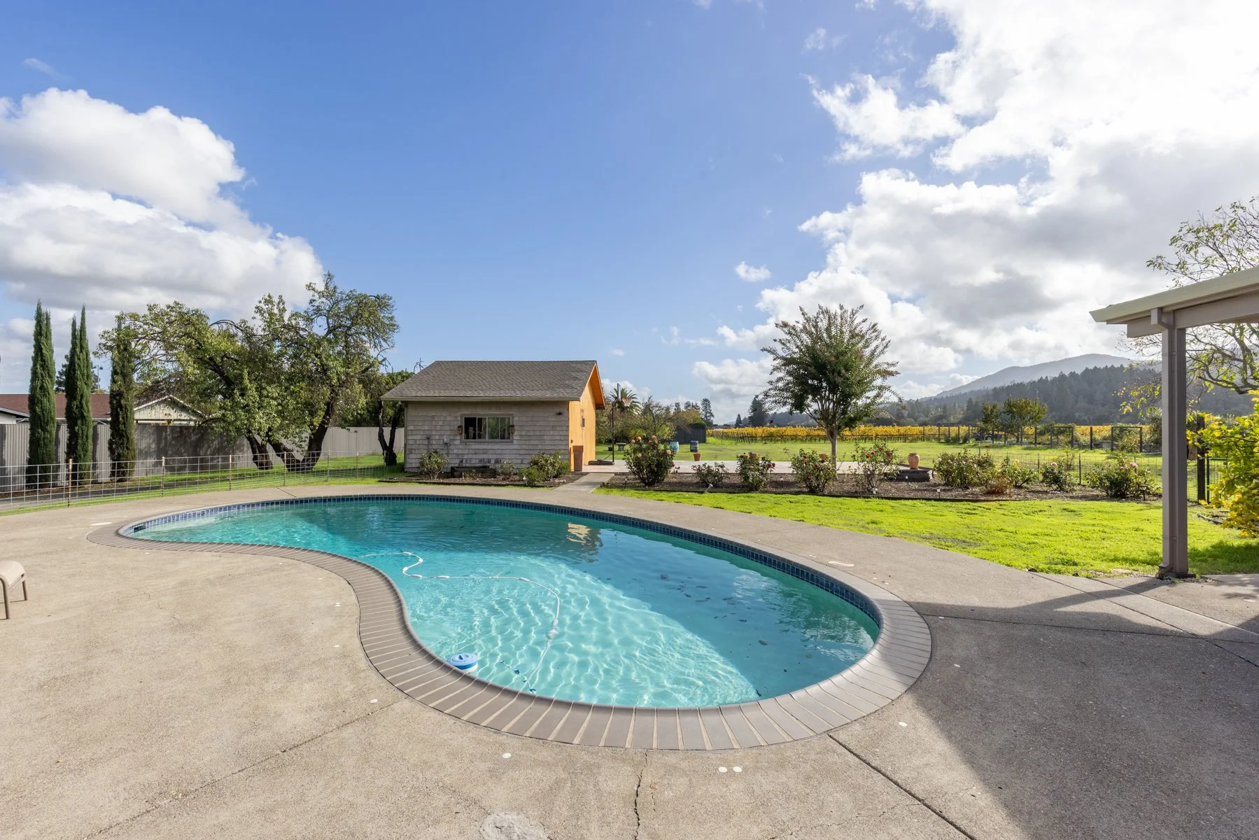 A backyard with a kidney-shaped swimming pool, surrounded by a concrete deck, with a small shed in the background and a scenic view of trees and mountains under a partly cloudy sky.