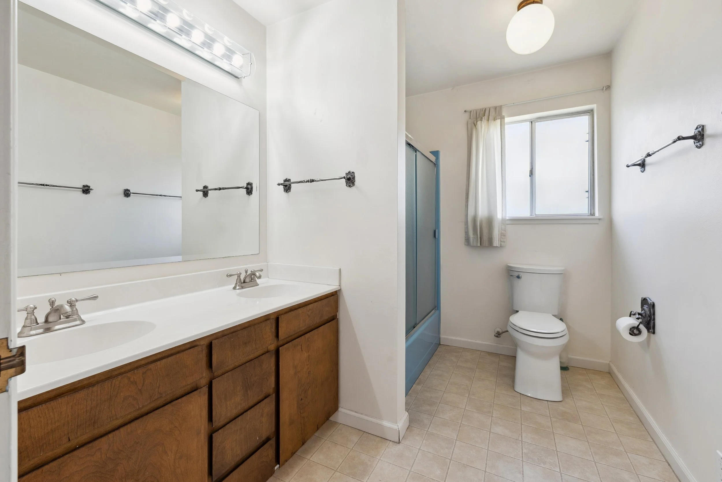 Empty bathroom with a double sink vanity, a mirror, a shower with blue trim, a toilet, and a window with a curtain.
