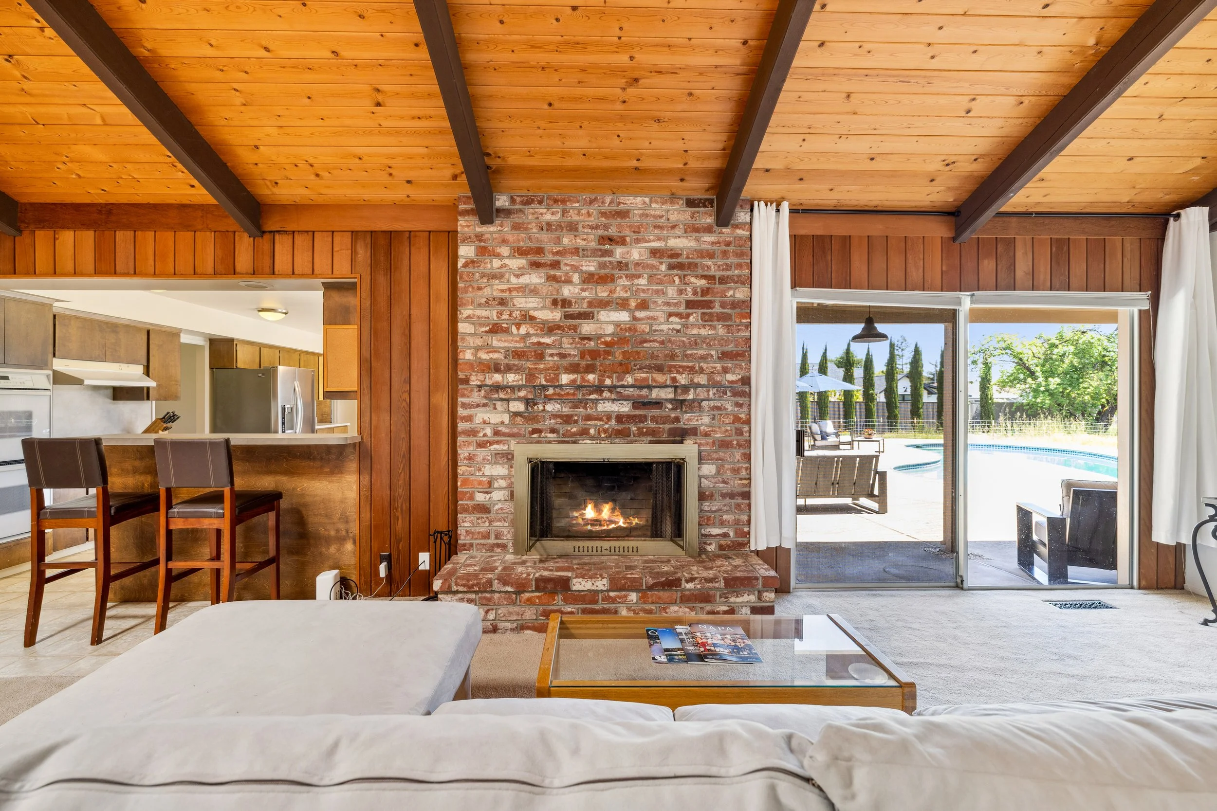 Living room with a brick fireplace, wooden ceiling, and sliding glass door leading to an outdoor patio with a swimming pool and outdoor furniture.
