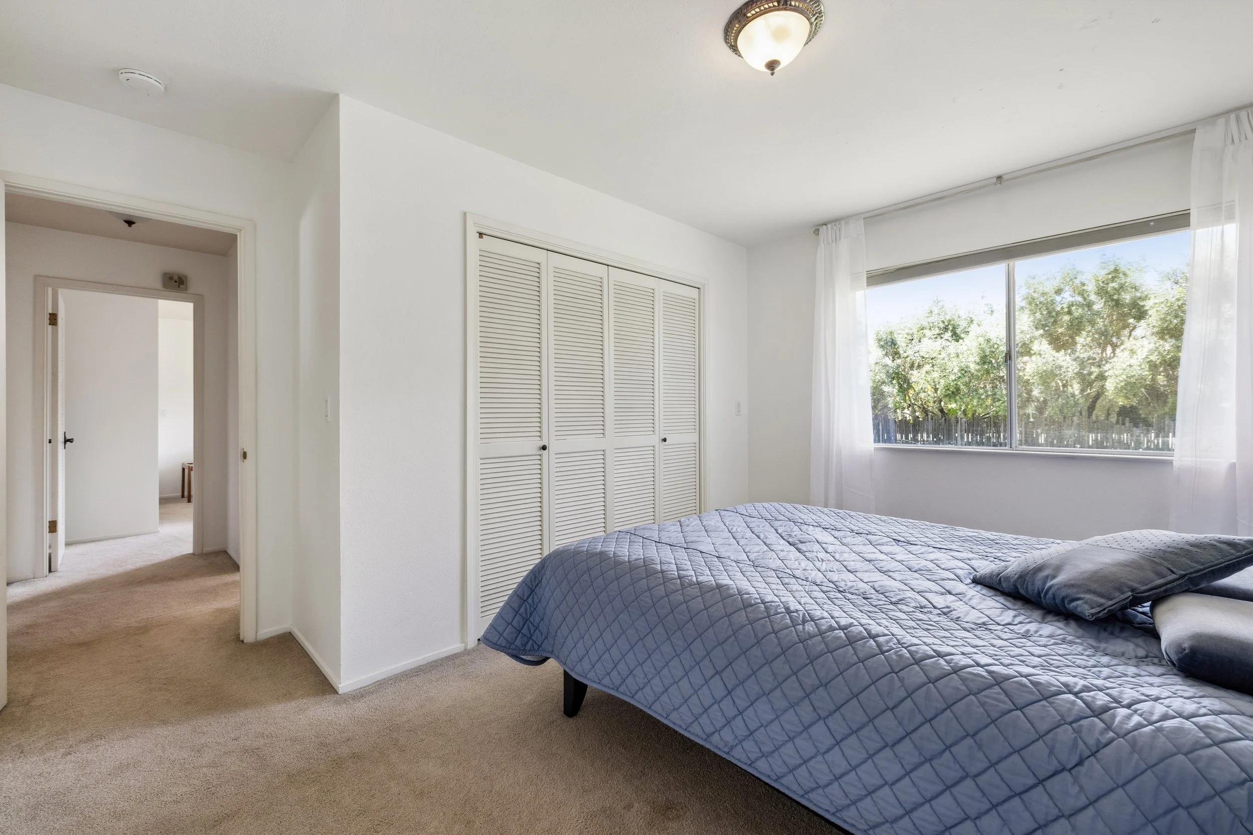 Bedroom with a bed covered with a blue quilt and pillows, a large window with white curtains showing green trees outside, a closet with louvered doors, and an entry door leading to a hallway.