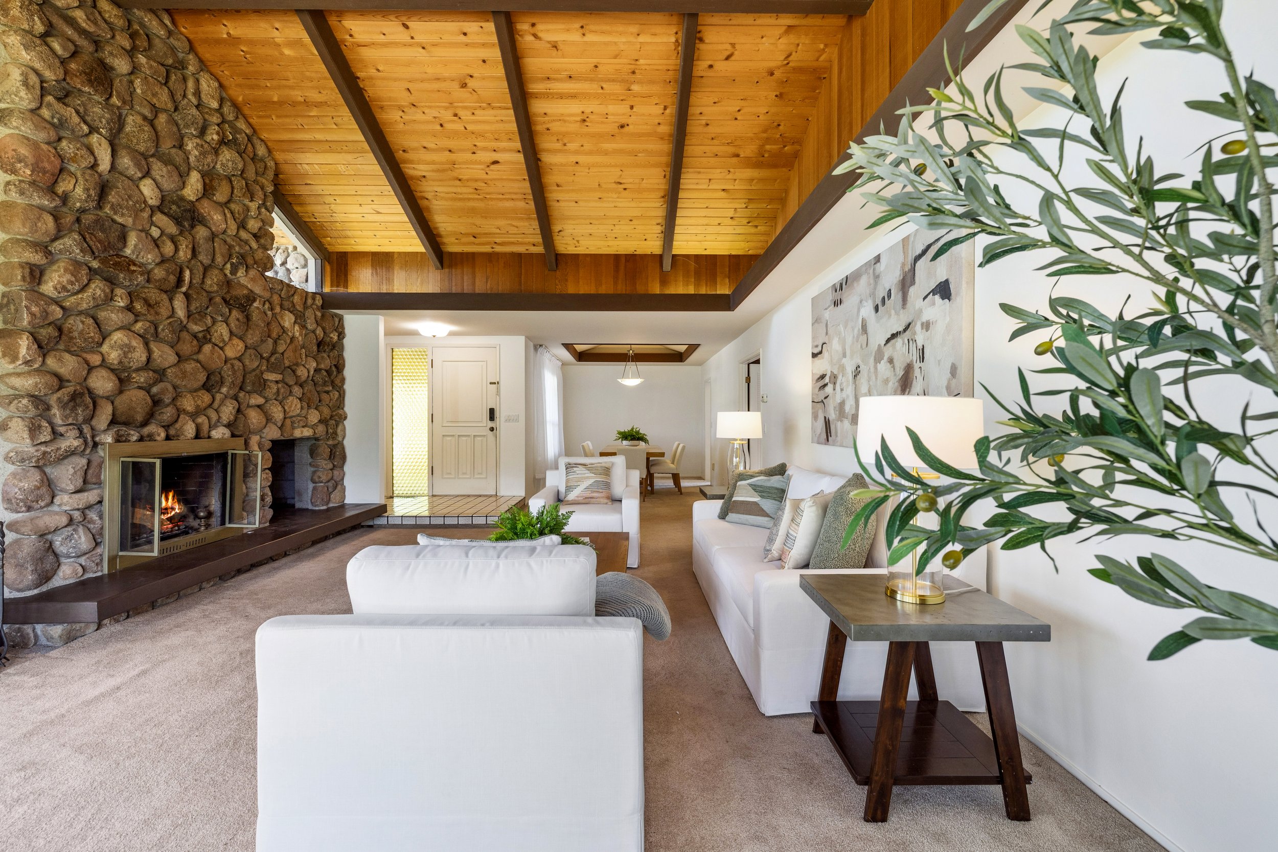 Living room with a stone fireplace, white sofas with pillows, a side table with a lamp, and a large plant on the right.