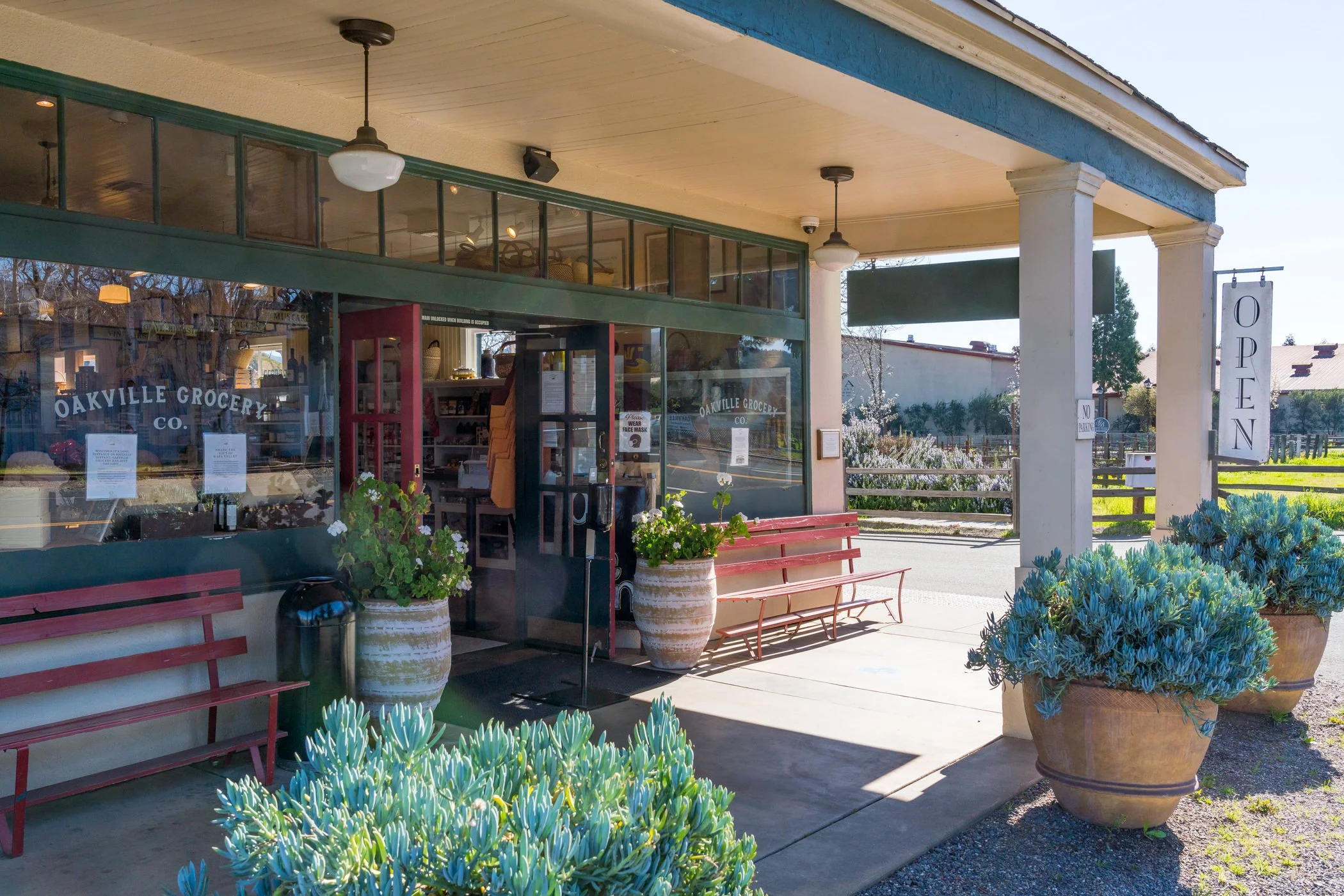 Exterior view of Oakville Grocery Co. with a glass storefront, red benches, large potted plants with greenery, and an 'Open' sign hanging on the porch. The scene is sunny with clear skies.