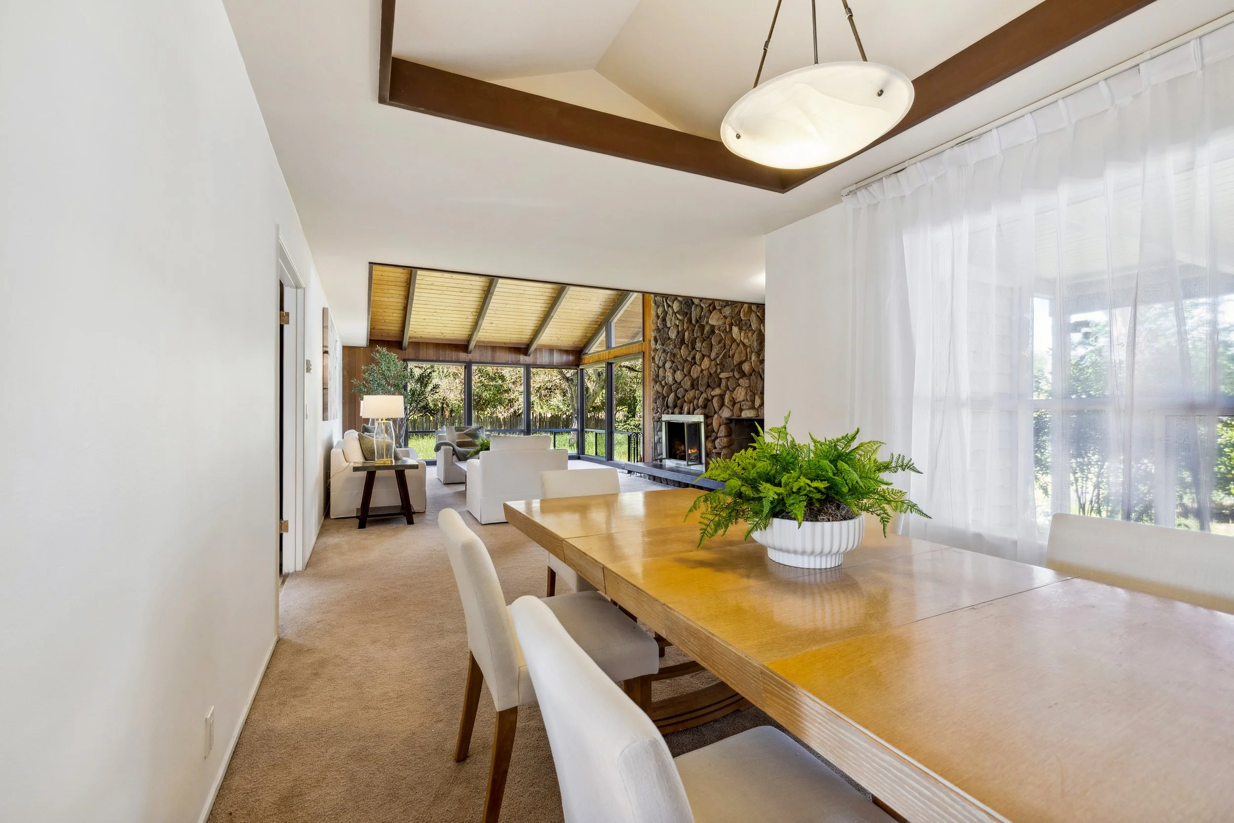 Open-concept living and dining room with a large wooden dining table, white chairs, and a potted fern. The space includes a stone fireplace, windows with sheer curtains, and a sitting area with white sofas and a side table with a lamp. The ceiling has a wooden beam and a modern light fixture.