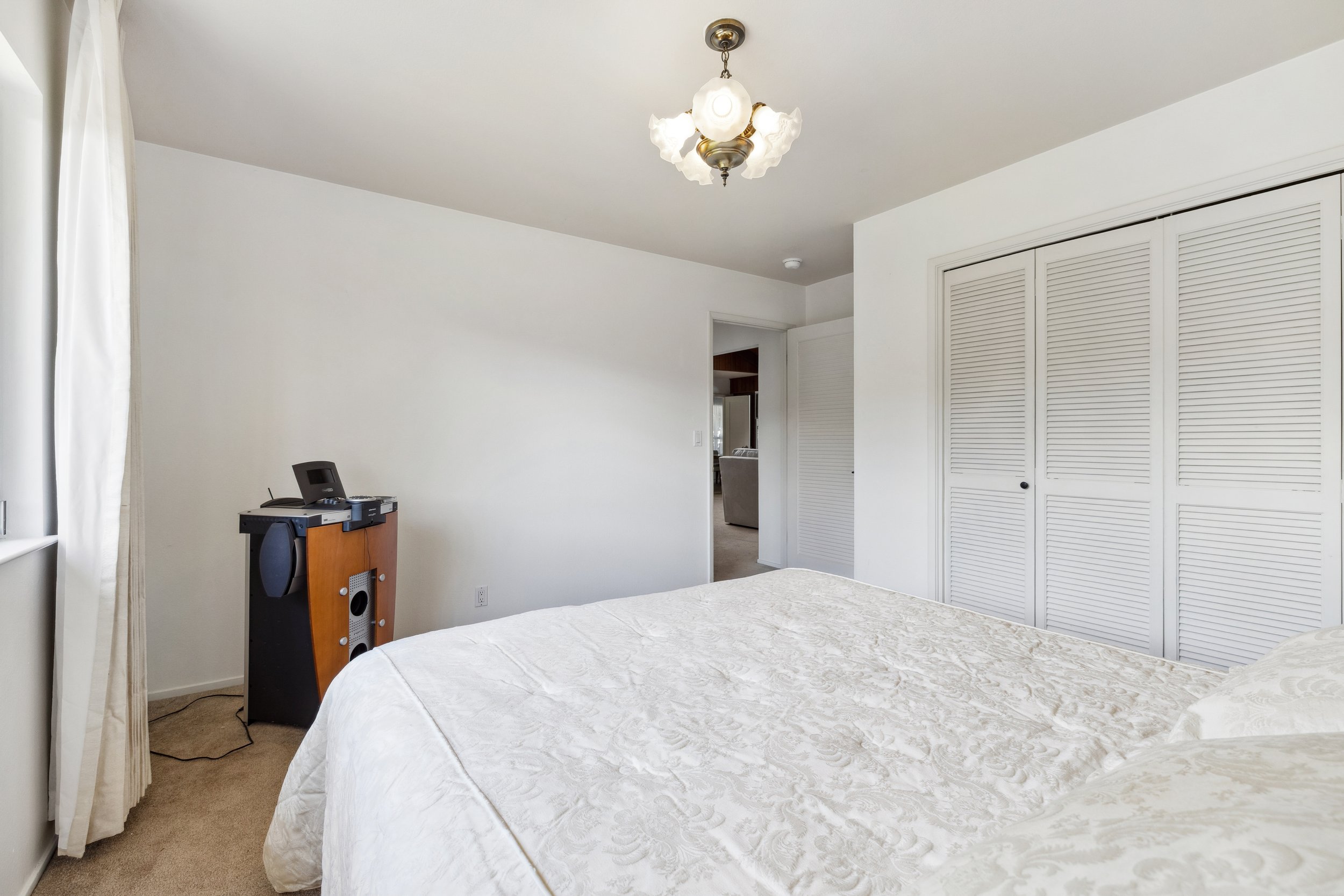 A bedroom with a bed covered in a white patterned bedspread. There is a closet with louvered doors on the right. An open doorway leads to another room with a couch. A small table with electronics is next to a window with white curtains. A ceiling lig