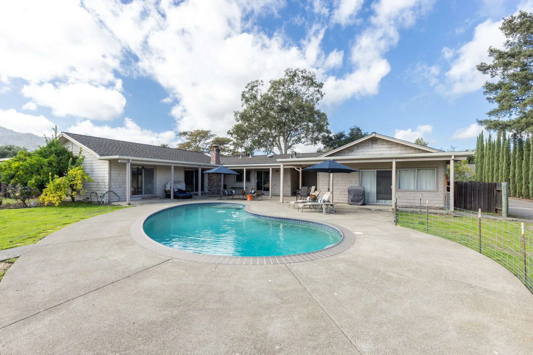 Backyard view featuring a kidney-shaped swimming pool, lounge chairs with umbrellas, a concrete patio, a single-story house with large windows, trees, and a partly cloudy sky.