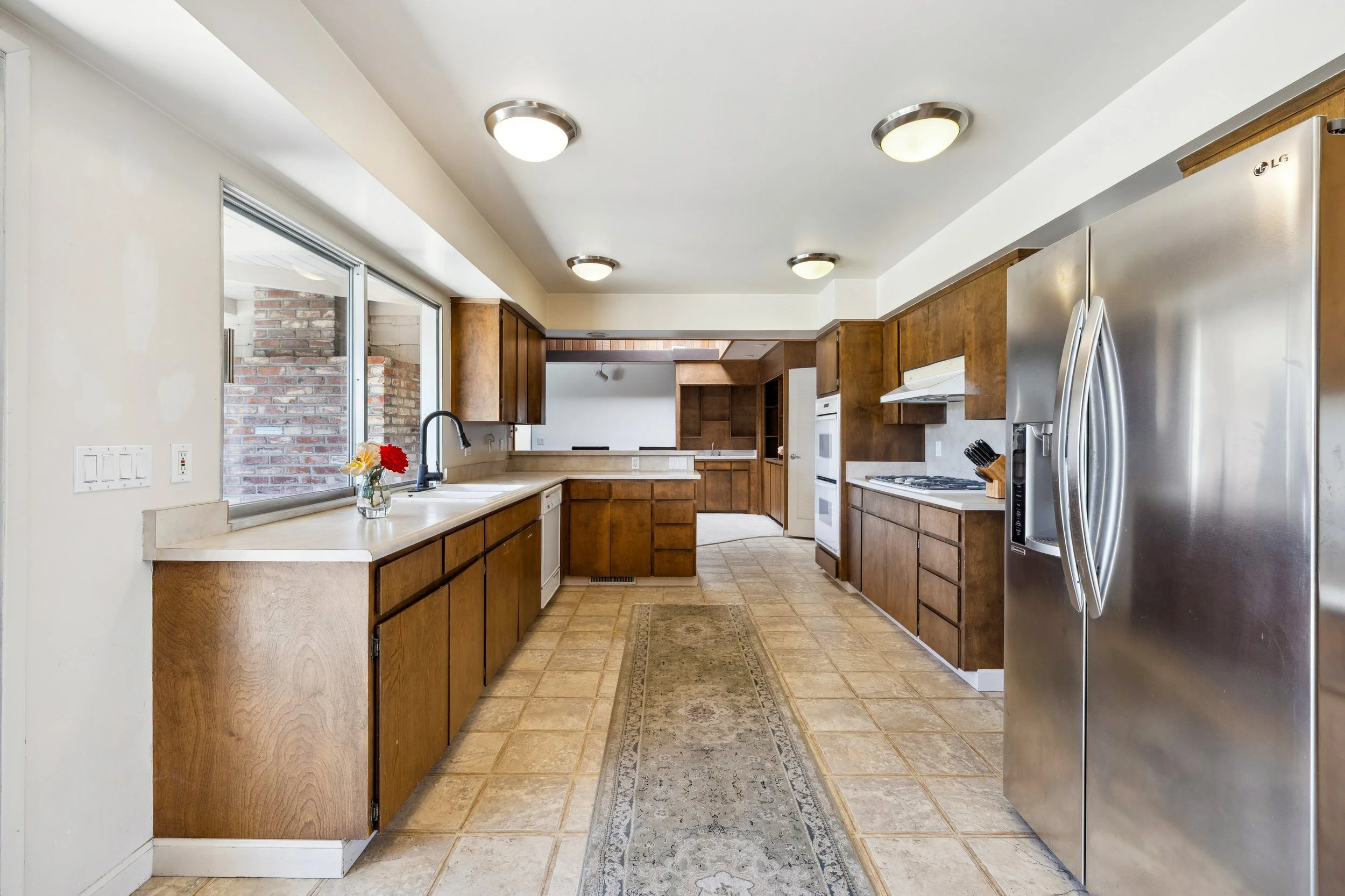 Kitchen with wooden cabinets, white countertops, stainless steel refrigerator, and a window overlooking a brick wall.