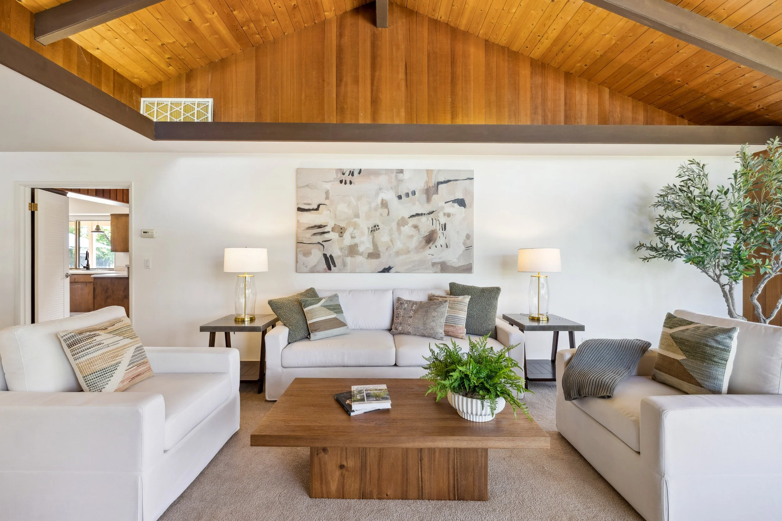 Living room with white sofas, a wooden coffee table with a plant, and gray and green cushions, with artwork on the wall, wooden ceiling, and a potted tree in the corner.