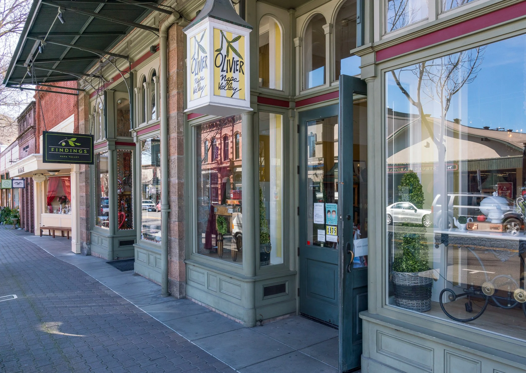 Exterior view of a storefront with large glass windows and a door, displaying signs for 'Findings Napa Valley' and 'Olivier Napa Valley,' with reflections of the street and parked cars visible in the windows.