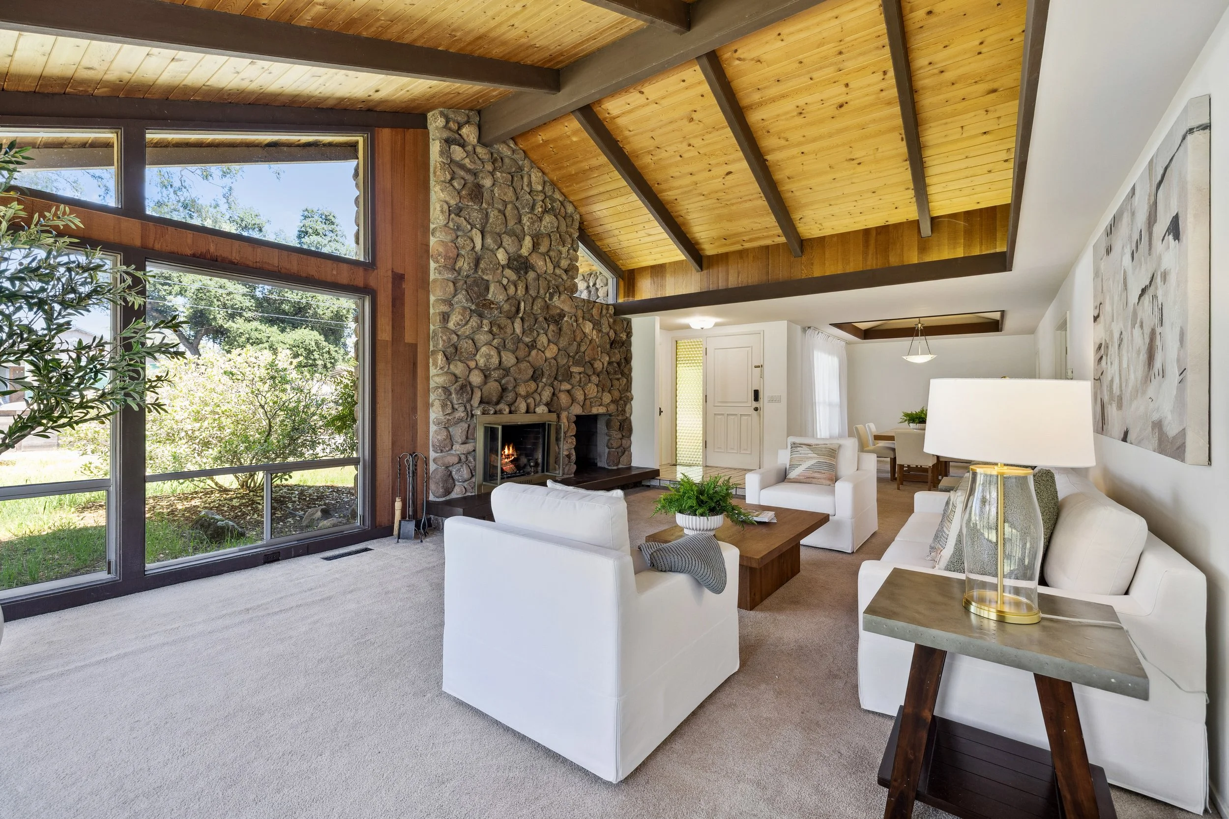 Living room with wood-paneled ceiling, stone fireplace, large windows, white furniture, and a view of outdoor greenery.