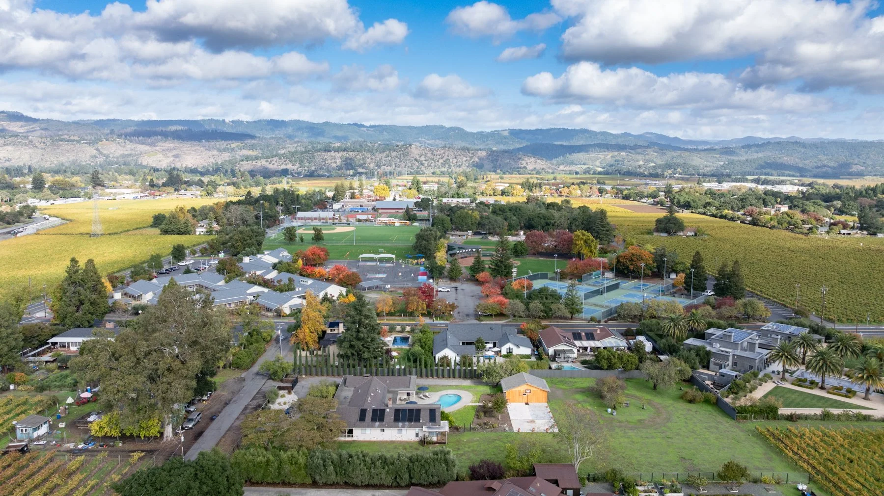 Aerial view of a small town and surrounding farmland with football and tennis courts, houses, trees, and mountains in the distance under a partly cloudy sky.