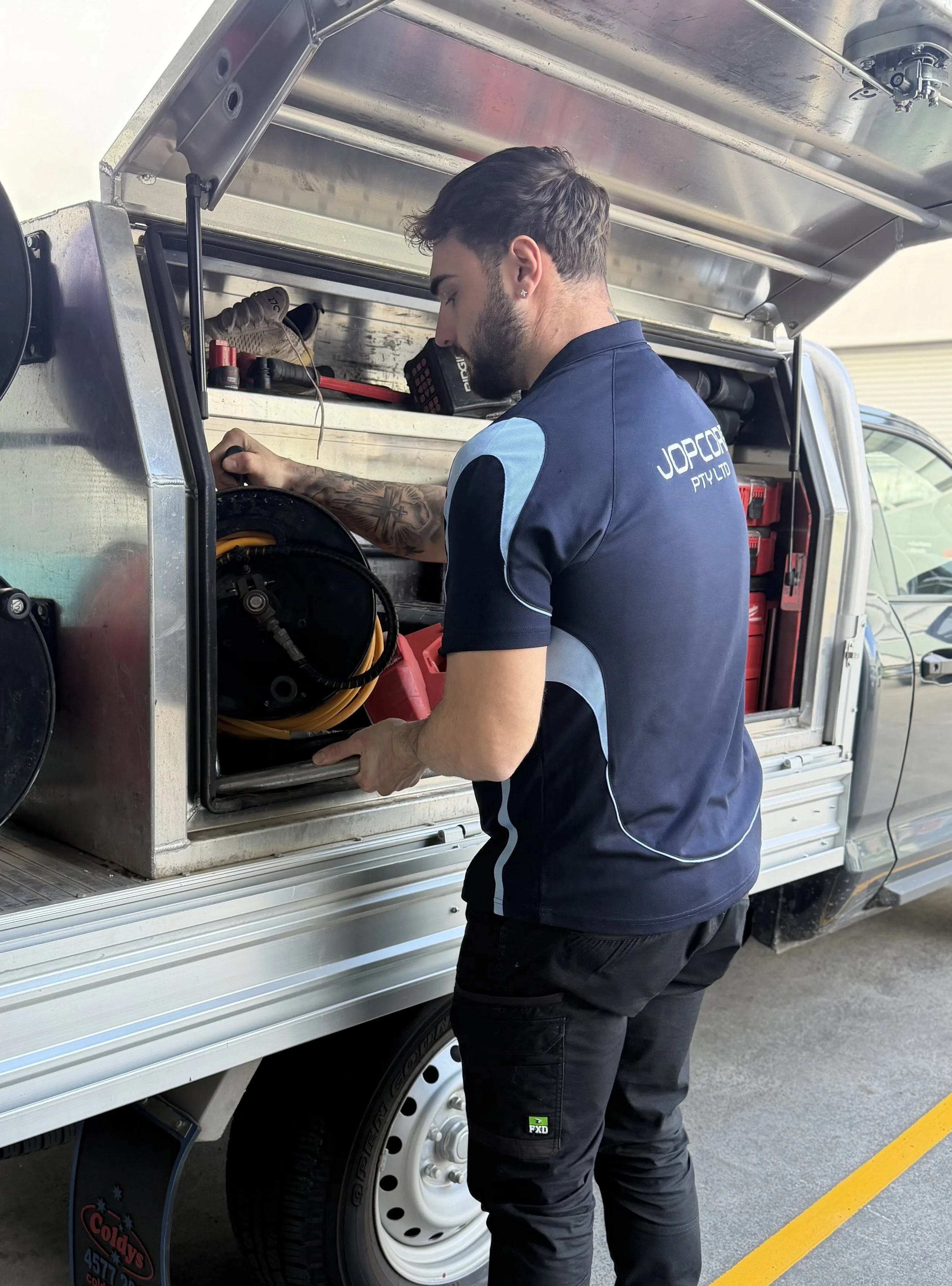 A man with a beard and tattooed arm working with equipment in the back of a service truck with an open side panel, wearing a dark blue polo shirt with light blue accents and the text 'JOPCON PTY LTD' on the back, in a parking lot marked with yellow lines.