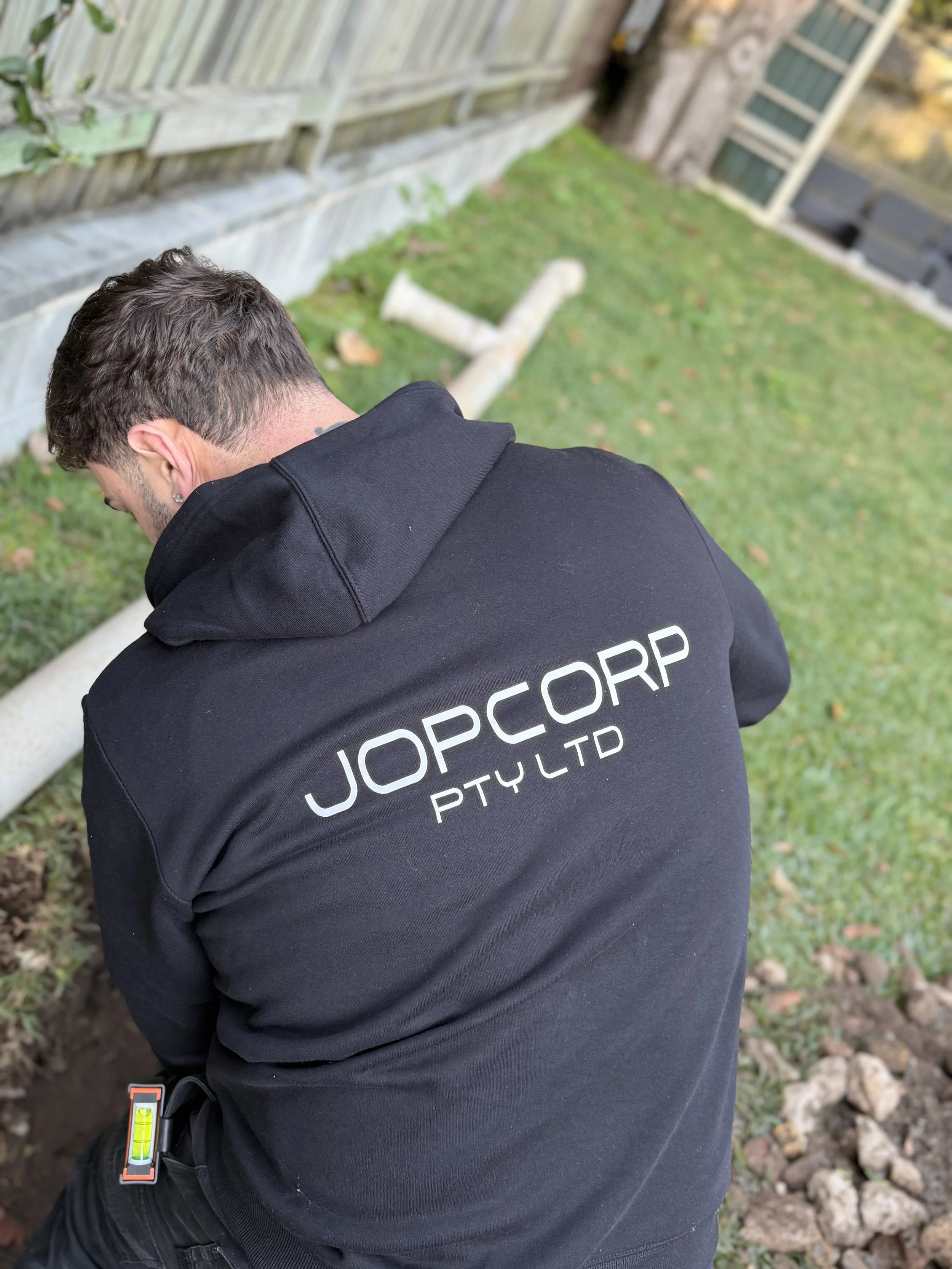 A person with short, dark hair wearing a black hoodie with white text on the back, kneeling or crouching near a garden bed with soil, laying next to a white pipe and a Spirit level tool, outdoors in a yard with a wooden fence and a house in the background.