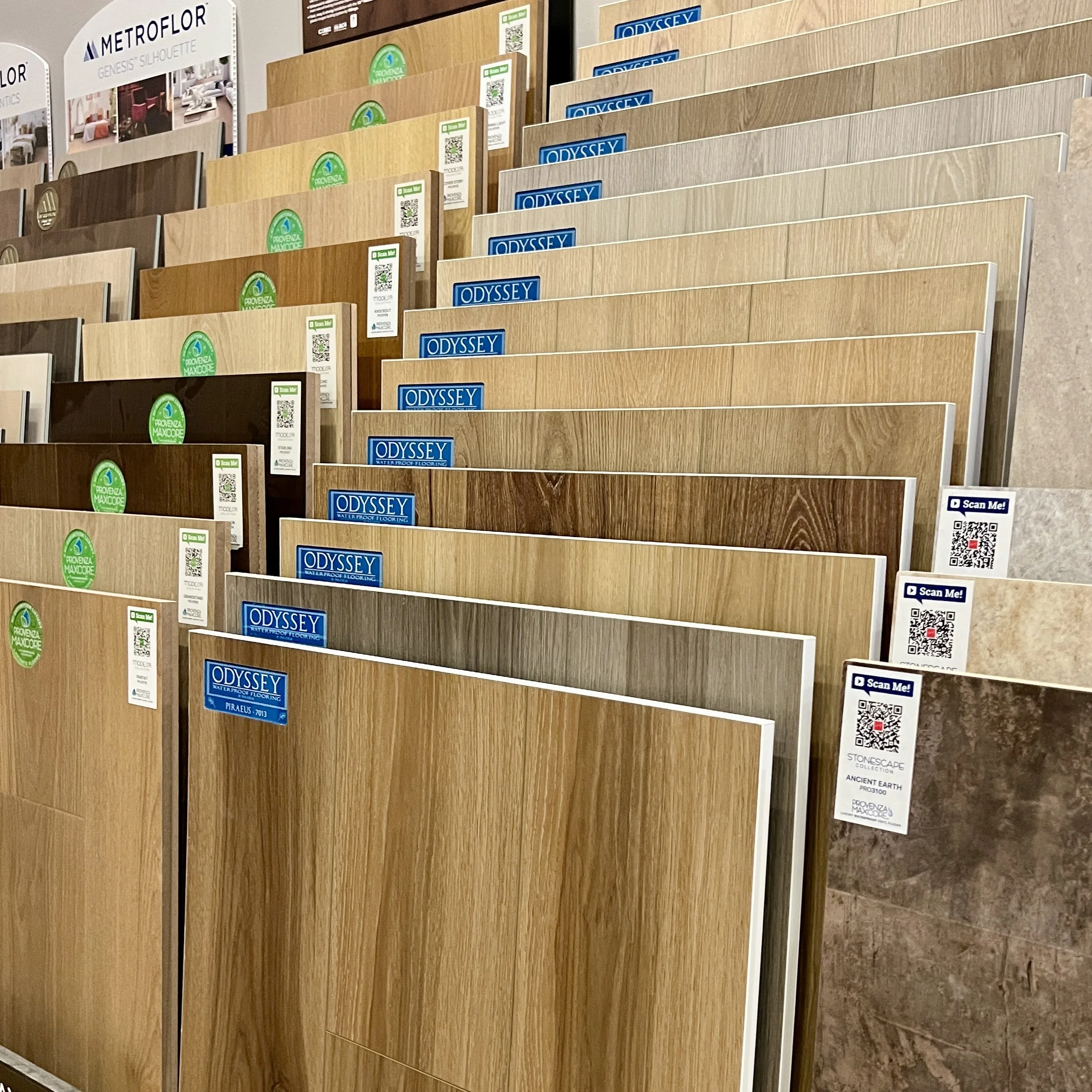Display of various luxury vinyl tile flooring sample boards, some labeled "Odyssey" and others labeled "Provenza Maxcore," arranged in a showroom setting.