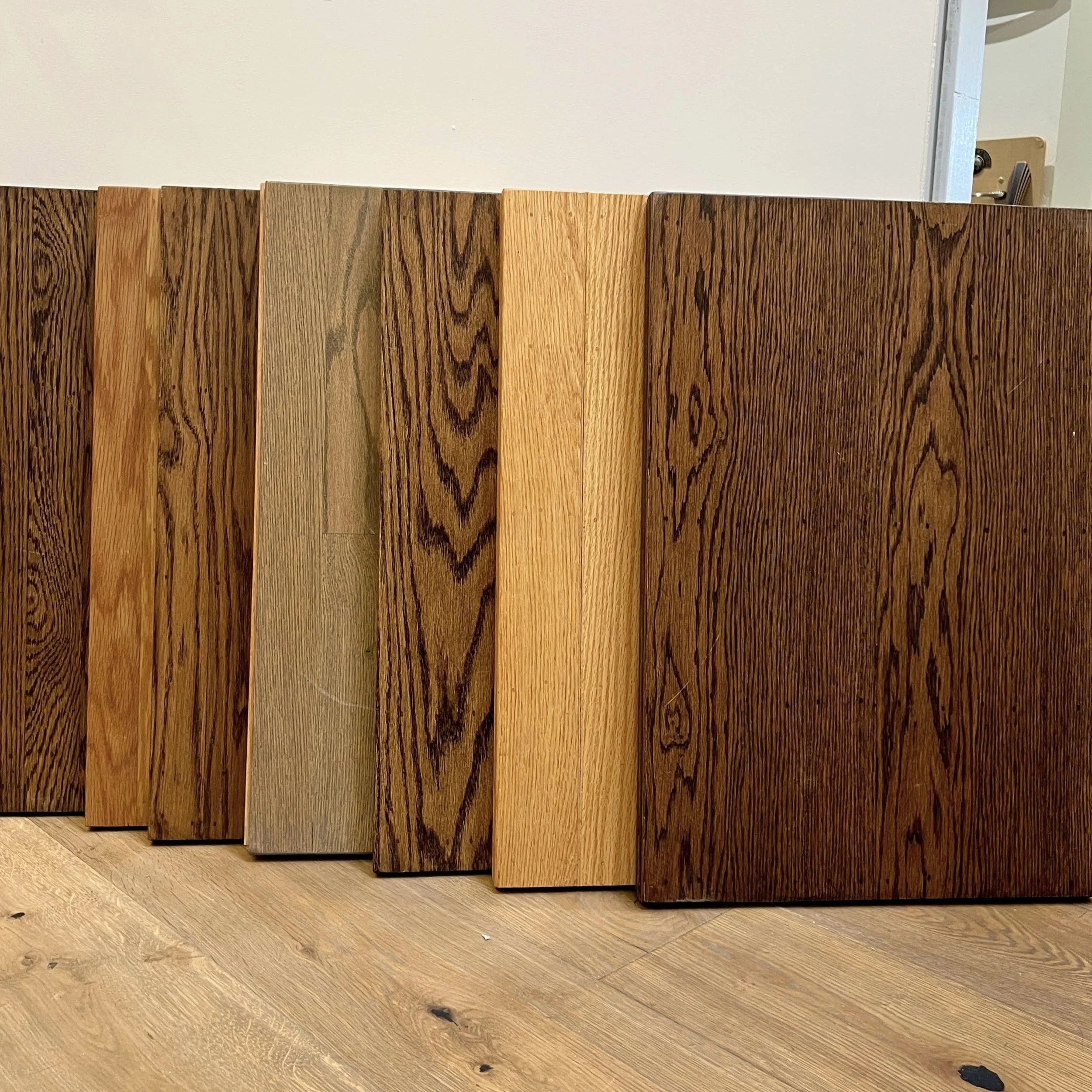 A row of solid oak hardwood flooring samples with varying shades of stains, standing upright on a light wood floor, with a white wall and a door in the background.