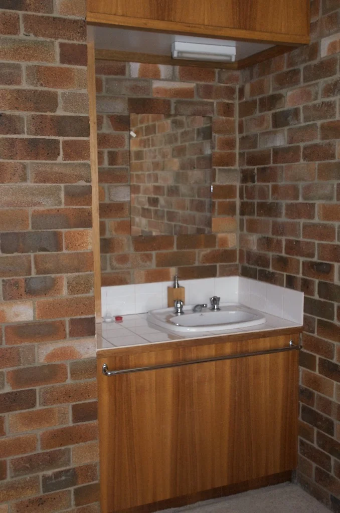 A small bathroom sink with a mirror above it, surrounded by brick walls and white tile backsplash, with a wooden cabinet below.